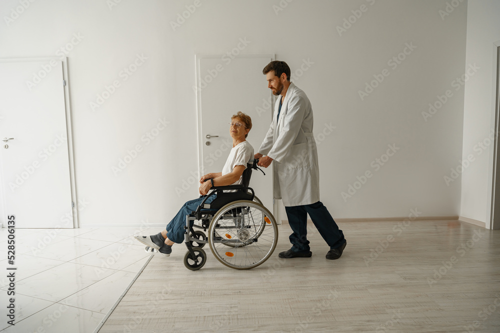 Professional male doctor carrying patient on wheelchair in medicine clinic hall. High quality photo Professional male doctor carrying patient on wheelchair in medicine clinic hall. High quality photo