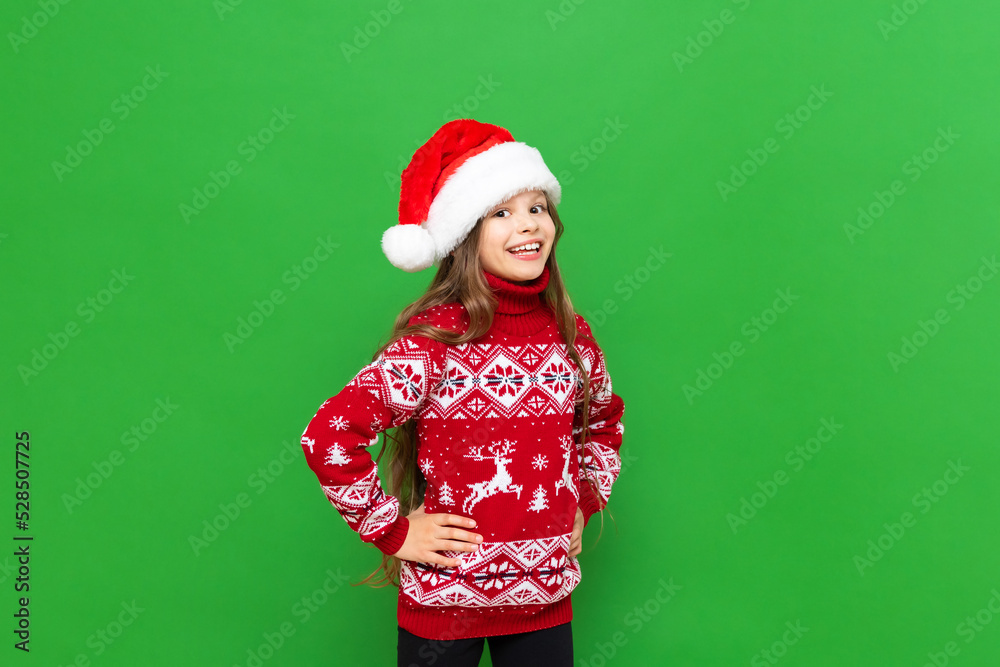 A little girl in a red sweater with reindeer and a Santa Claus hat waiting for the holiday on a green isolated background. The concept of Christmas. A little girl in a red sweater with reindeer and a Santa Claus hat waiting for the holiday on a green isolated background. The concept of Christmas.