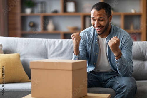Happy black man looking at cardboard box at home and celebrating success © Prostock-studio