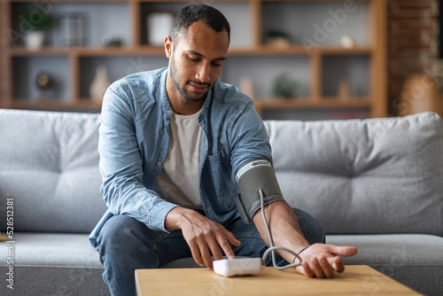 Fototapeta Black Man Sitting On Couch, Checking Blood Pressure With Upper Arm Monitor