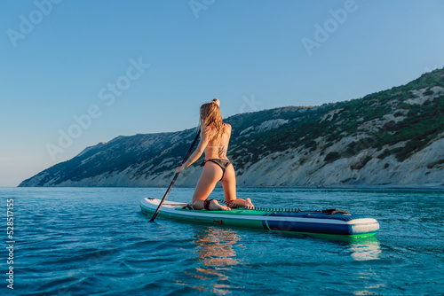 Bikini girl rowing on stand up paddle board at sea. Woman on SUP board in sea with mountain coastline. © artifirsov