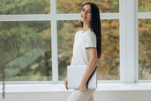 Free Download A young woman stands near a window with a view of nature and holds a closed laptop in her hands - GFXLoad