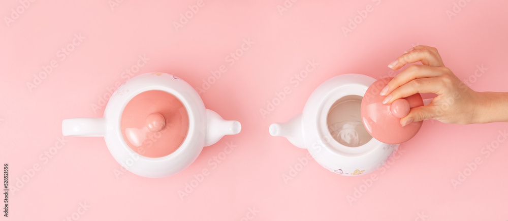 Banner. Two white porcelain teapots with a pink lid on a pink background. Female hand removes the lid from the kettle. Flat lay. Copy space. Concept of Breast Cancer Awareness Month - obrazy, fototapety, plakaty 