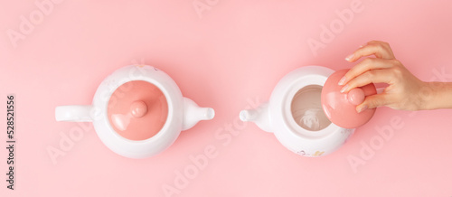Banner. Two white porcelain teapots with a pink lid on a pink background. Female hand removes the lid from the kettle. Flat lay. Copy space. Concept of Breast Cancer Awareness Month