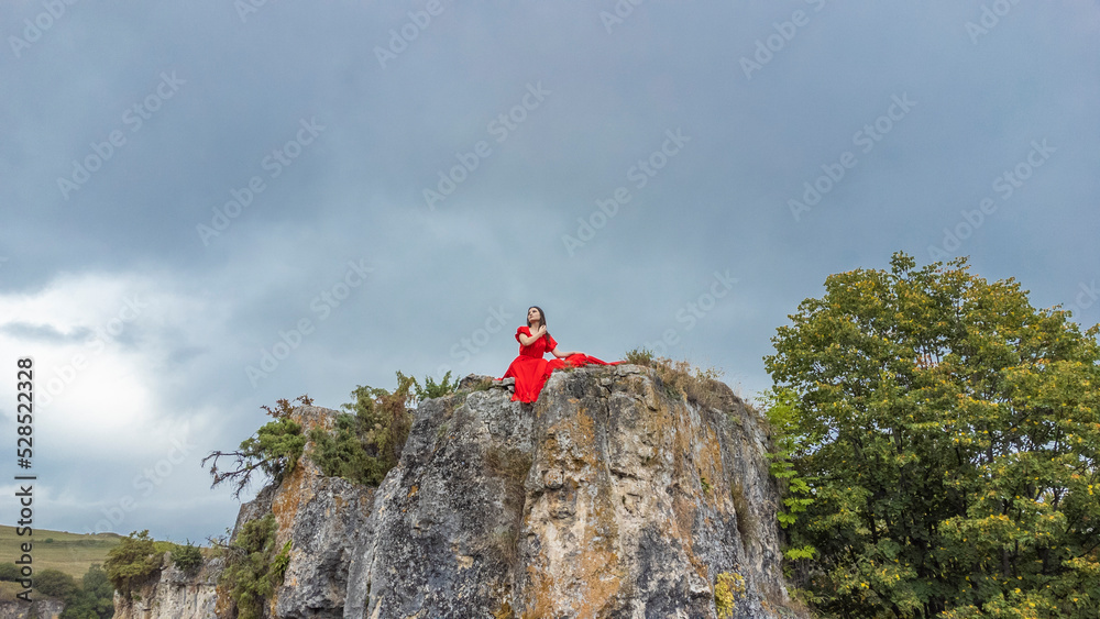 A girl in a red dress stands on the edge of a cliff, summer, mountains and forest, a seed from a drone