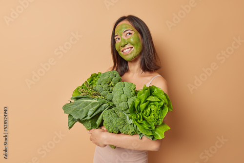 Pretty smiling Asian woman with dark hair applies beauty green mask carries fresh vegetables has dreamy expression imagines something dressed in sportswear has slim figure isolated over beige wall