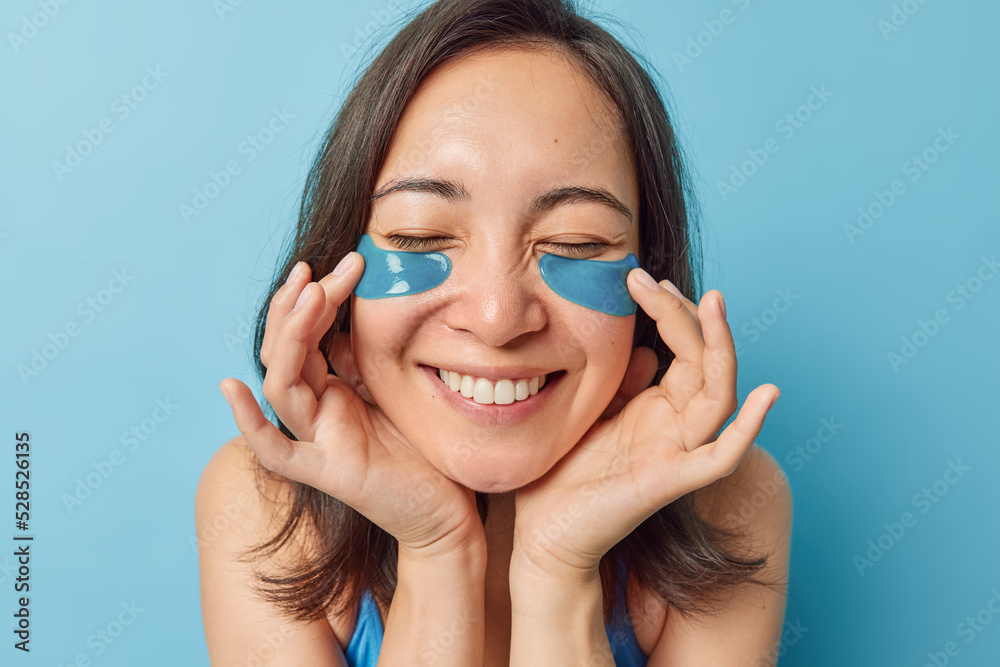 Headshot of beautiful happy dark haired woman applies hydrogel beauty patches under eyes to reduce wrinkles smiles toothily has healthy clean skin undergoes facial treatments isolated on blue wall