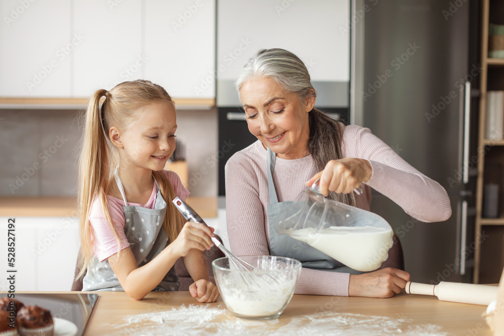 Happy european elderly grandmother and little granddaughter in aprons pour milk to dough