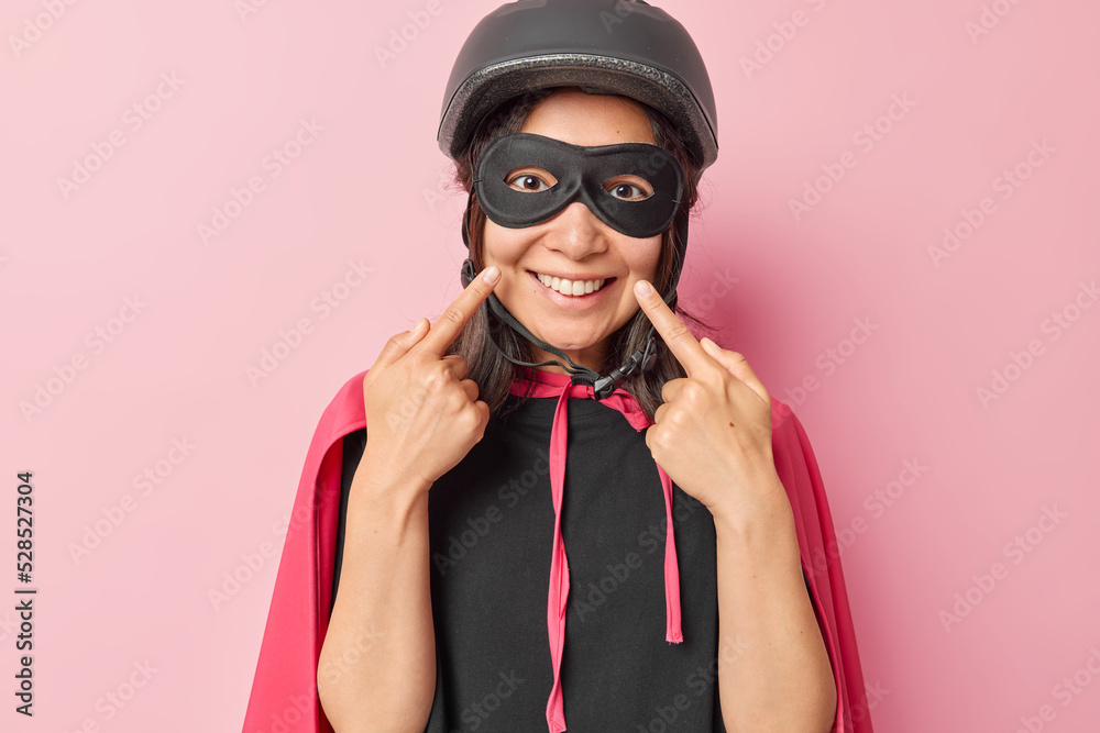 Horizontal shot of cheerful woman dressed in superhero costume points at toothy smile with index fingers has happy face wears helmet eyemask and cape isolated over pink background. Look at my teeth Horizontal shot of cheerful woman dressed in superhero costume points at toothy smile with index fingers has happy face wears helmet eyemask and cape isolated over pink background. Look at my teeth