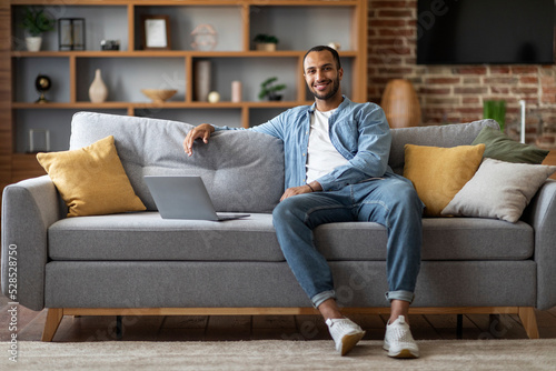 Handsome Black Man Sitting On Couch With Laptop And Smiling At Camera