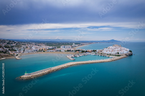 Fotografía de la ciudad amurallada de Peñíscola y su puerto pesquero al completo con el cielo parcialmente nublado.