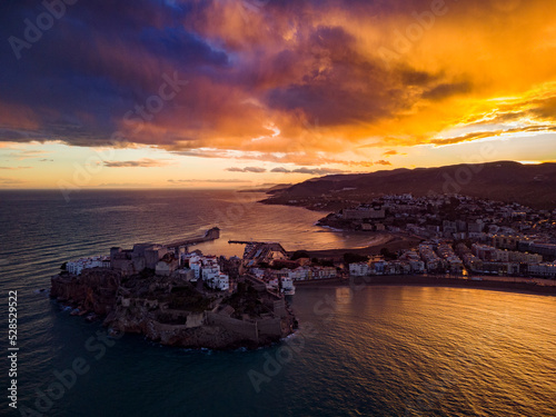 Fotografía aérea de la ciudad amurallada de Peñíscola y su castillo al atardecer con el cielo nublado y colores de fuego.