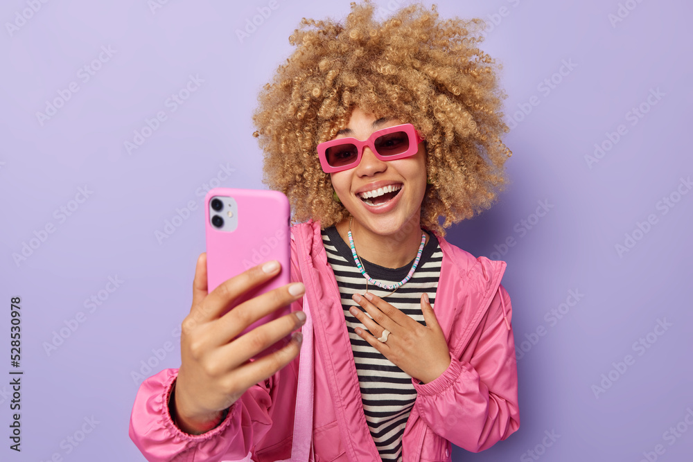 Positive curly haired young European woman wears trendy sunglasses striped jumper and jacket takes selfie makes video call has happy conversation with best friend isolated over purple background. Positive curly haired young European woman wears trendy sunglasses striped jumper and jacket takes selfie makes video call has happy conversation with best friend isolated over purple background.