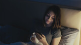 Online connection. Sleepless woman. Pretty lady in bed scrolling social media in smartphone drinking tea in light shadow room interior.