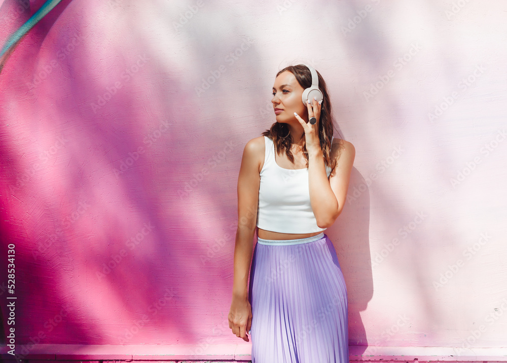 Obraz premium a young excited woman in a top and skirt, wearing headphones, listening to music, walking, dancing with her hands up in the open air against a pink wall.