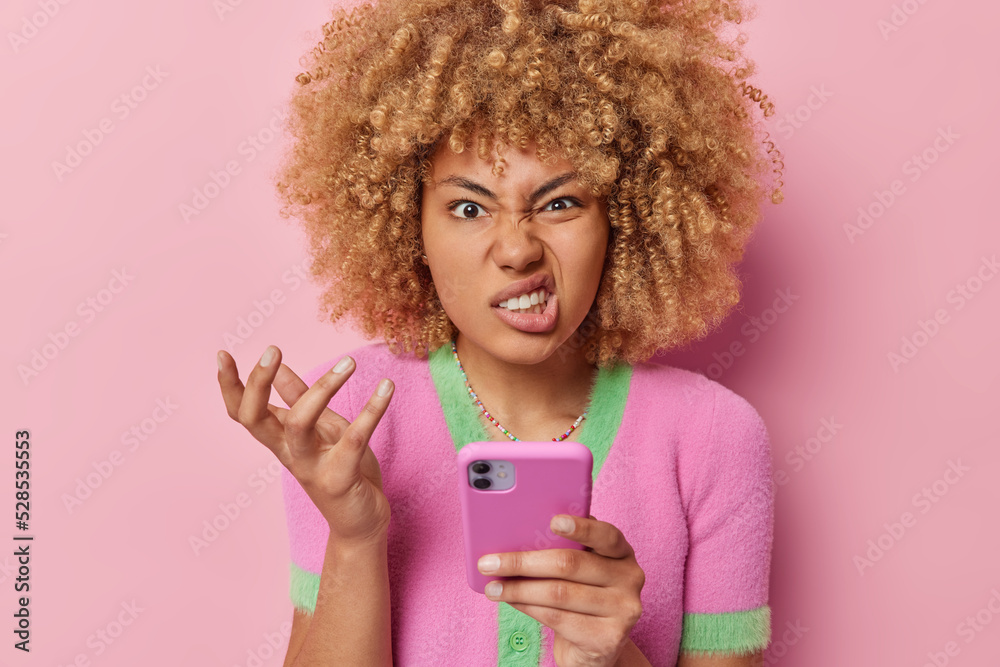 Annoyed curly haired woman holds mobile phone stares furious and ...