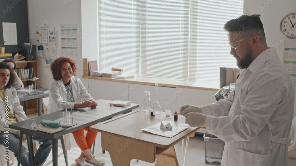 Side view of male chemistry teacher in medical gown and protective goggles standing in front of high school students in morning, making experiment with chemicals Side view of male chemistry teacher in medical gown and protective goggles standing in front of high school students in morning, making experiment with chemicals