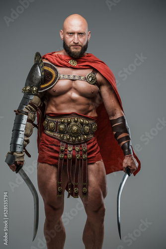 Portrait of greek gladiator from past dressed in armor holding swords isolated on grey.