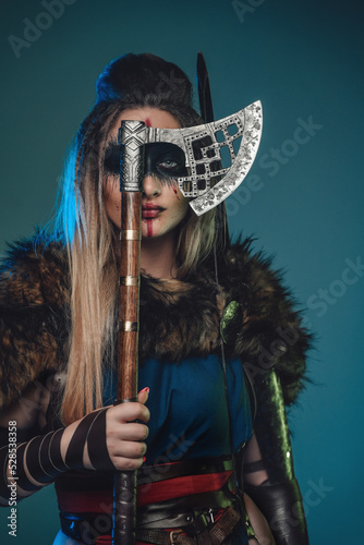 Portrait of ancient valkyrie with fur coat hiding her face with hatchet and looking at camera.