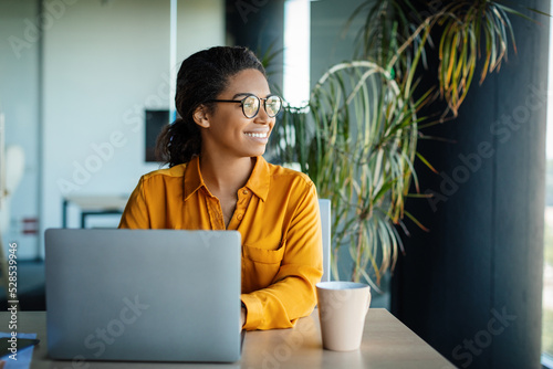 Happy dreamy woman sitting at workplace with laptop and looking away with glad face expression, copy space photo