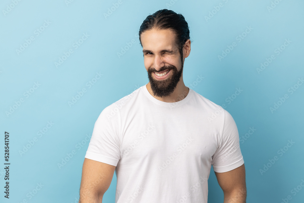 Portrait of cheerful man with beard wearing white T-shirt being in good mood, smiling broadly and winking at camera with toothy smile. Indoor studio shot isolated on blue background. Portrait of cheerful man with beard wearing white T-shirt being in good mood, smiling broadly and winking at camera with toothy smile. Indoor studio shot isolated on blue background.