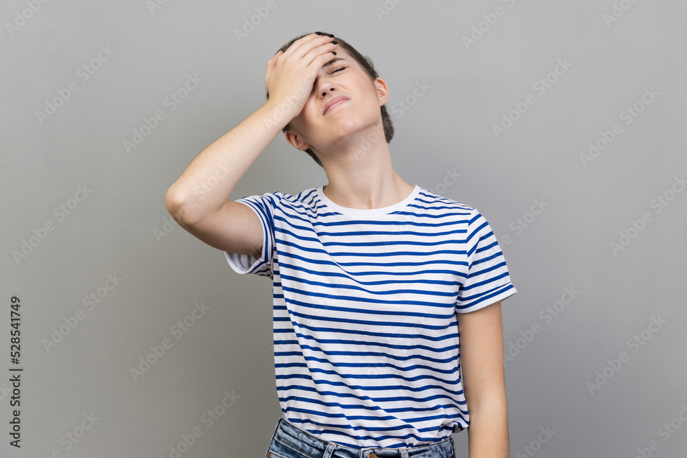 Portrait of upset frustrated woman wearing striped T-shirt holding forehead with hand, making facepalm gesture, forget about date, big failure. Indoor studio shot isolated on gray background. Portrait of upset frustrated woman wearing striped T-shirt holding forehead with hand, making facepalm gesture, forget about date, big failure. Indoor studio shot isolated on gray background.