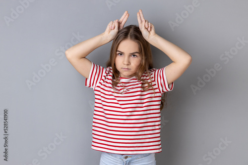 Portrait of aggressive bully little girl wearing striped T-shirt showing bull horns gesture over head, frowning as before attack. Indoor studio shot isolated on gray background.