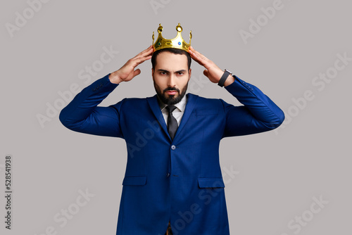 Serious independent bearded man wearing golden crown, looking with arrogance and confidence, privileged status, wearing official style suit. Indoor studio shot isolated on gray background. photo