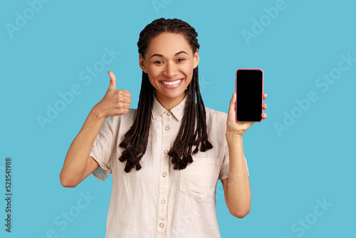 Joyful woman with black dreadlocks smiles happily, points at blank screen of cellualr, advertises new gadget, showing thumb up, wearing white shirt. Indoor studio shot isolated on blue background.