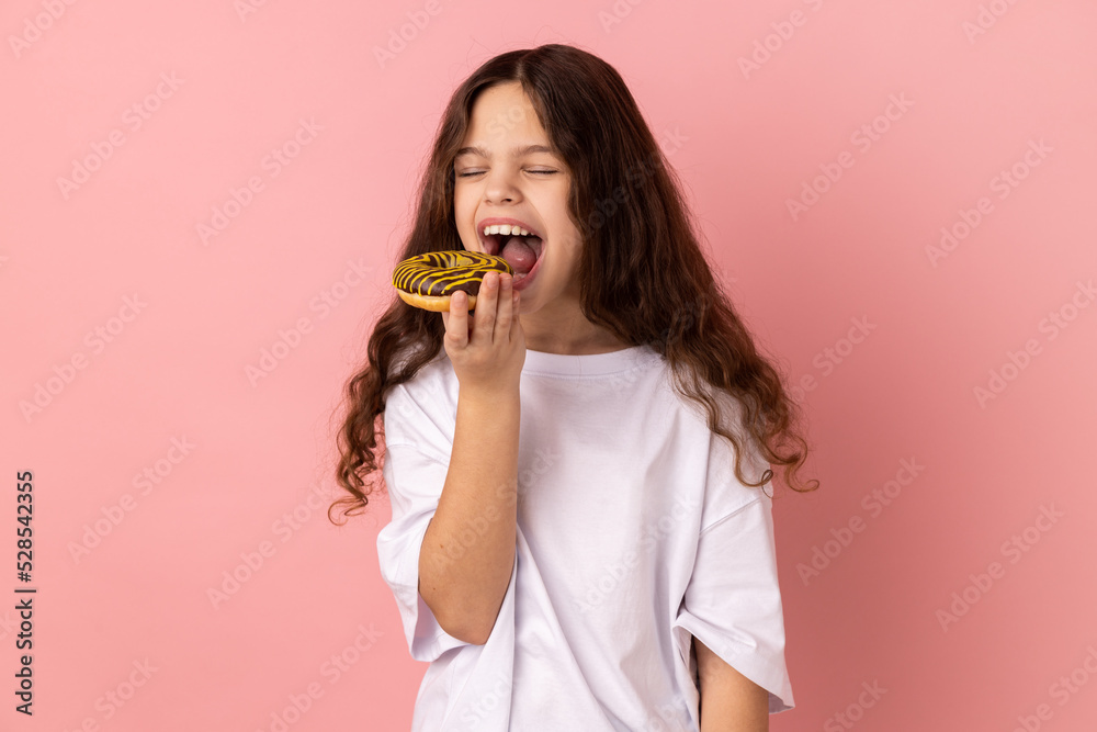 Sugar addiction. Portrait of satisfied little girl wearing white T-shirt biting delicious donut, looking with desire to eat sweet dessert. Indoor studio shot isolated on pink background. Sugar addiction. Portrait of satisfied little girl wearing white T-shirt biting delicious donut, looking with desire to eat sweet dessert. Indoor studio shot isolated on pink background.