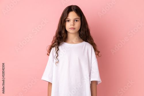 Portrait of serious strict little girl wearing white T-shirt standing looking at camera with bossy expression, being in bad mood. Indoor studio shot isolated on pink background.