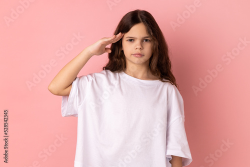 Fotobehang Portrait of serious little girl wearing white T-shirt giving salute, obediently listening to commander order with attentive confident face