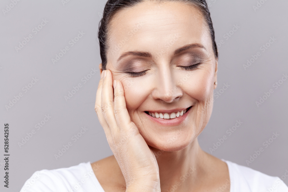 Closeup portrait of middle aged woman gently touching face with one hand and smiling, having healthy pure skin, female enjoying spa facial treatment. Indoor studio shot isolated on gray background. Closeup portrait of middle aged woman gently touching face with one hand and smiling, having healthy pure skin, female enjoying spa facial treatment. Indoor studio shot isolated on gray background.
