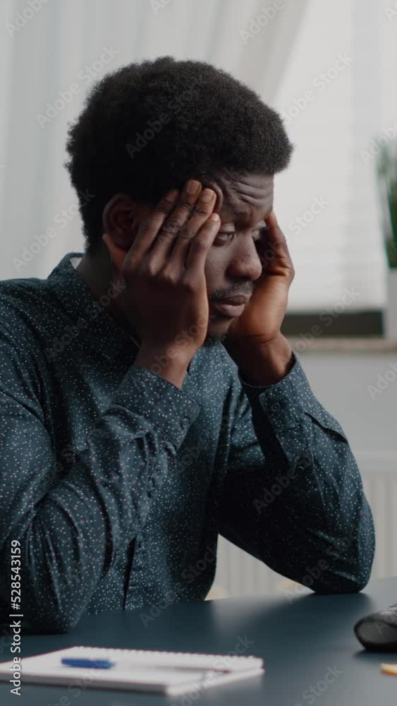 Vertical video: Stressing african american man with big headache pain, suffering from migraine, rubbing his temples. Overwhelmed manager working from home office, exhausted guy from problems and job Vertical video: Stressing african american man with big headache pain, suffering from migraine, rubbing his temples. Overwhelmed manager working from home office, exhausted guy from problems and job