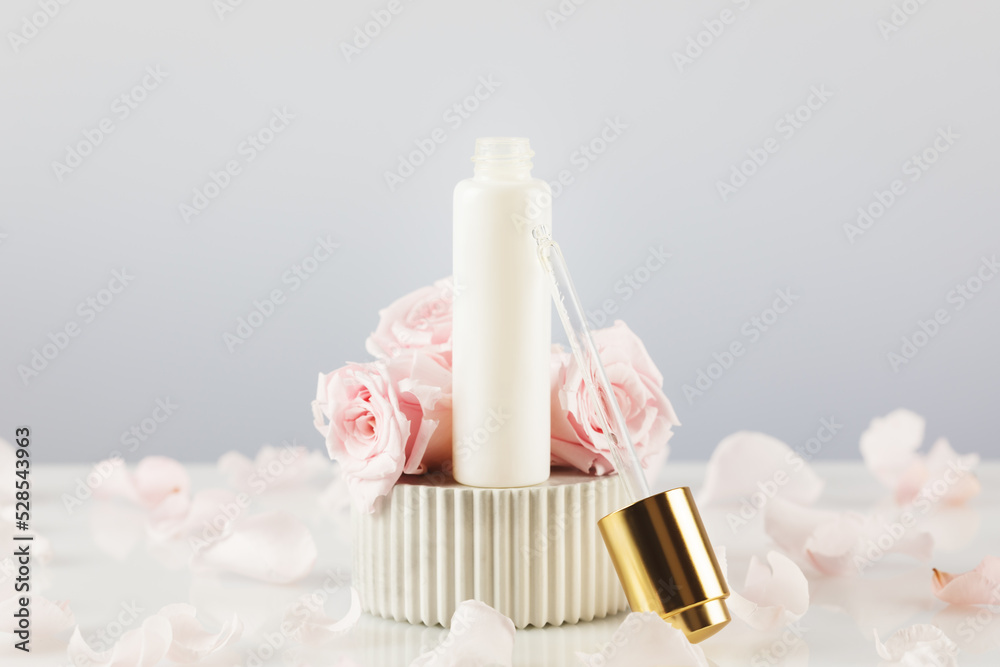 White dropper bottle with serum on a marble podium on a white table with tender pink roses and rose petals. Natural beauty product based on rose flowers. Soft focus style White dropper bottle with serum on a marble podium on a white table with tender pink roses and rose petals. Natural beauty product based on rose flowers. Soft focus style