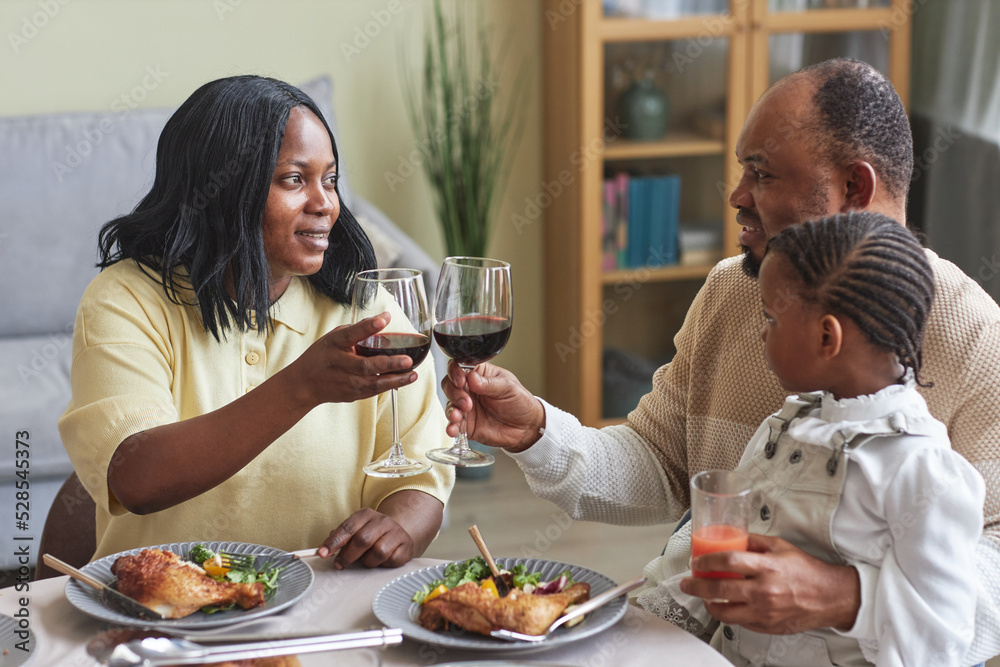 African couple toasting with glasses of red wine while having dinner with child together at dining table African couple toasting with glasses of red wine while having dinner with child together at dining table