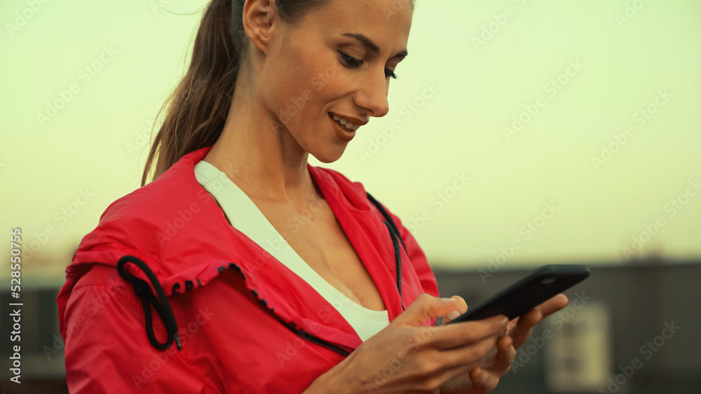 Naklejka premium Positive brunette sportswoman using mobile phone on blurred roof in evening.