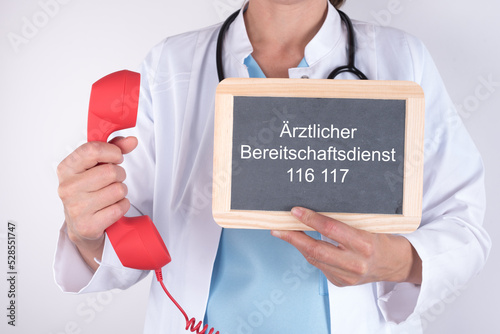 Ärztin mit einem roten Notfalltelefon und einer Tafel auf der Ärztlicher Bereitschaftsdienst 116 117 steht photo