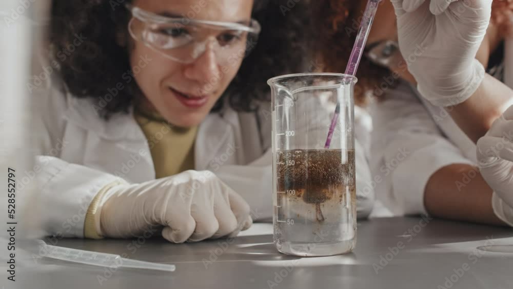 Low angle midsection of two students in white gowns and gloves sitting at school desk, watching chemical reaction in beaker at daytime, talking Low angle midsection of two students in white gowns and gloves sitting at school desk, watching chemical reaction in beaker at daytime, talking