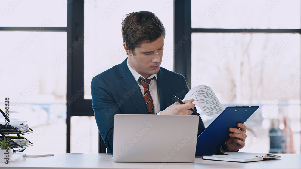 serious businessman looking at document near laptop in office. serious businessman looking at document near laptop in office.