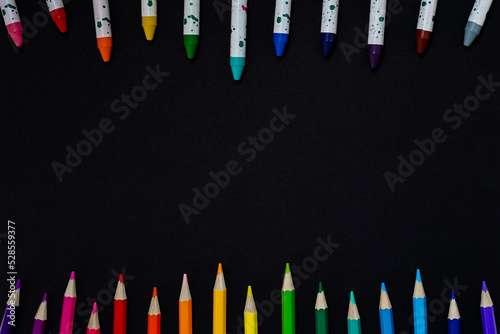 Frame of colored pencils and wax crayons, black background with space in the middle for ad text. Multicolored pencils, crayons laid out in rainbow colors on the top and bottom edges of the blackboard.