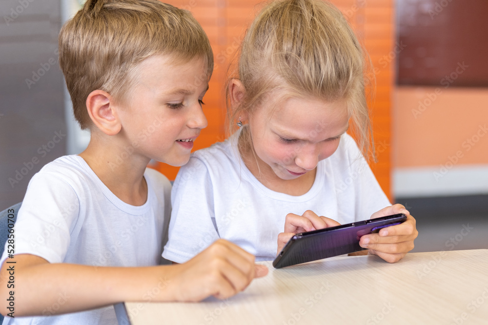 A little girl and a boy are watching cartoons on their phone, sitting at a table. Children play an online game, communicate virtually using the phone. Education, lifestyle A little girl and a boy are watching cartoons on their phone, sitting at a table. Children play an online game, communicate virtually using the phone. Education, lifestyle