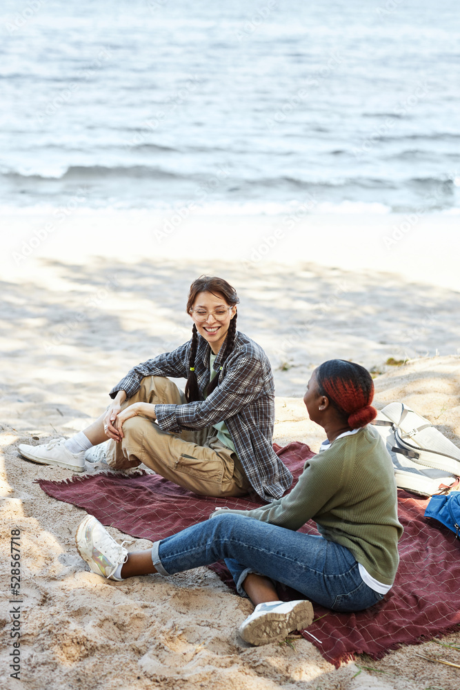Two young girls sitting on seashore and talking to each other during picnic outdoors