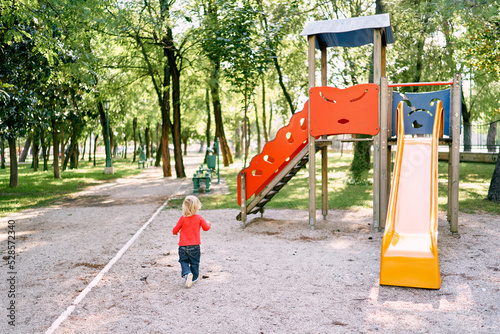 Little girl runs through the green park to the slide on the playground. Back view. High quality photo