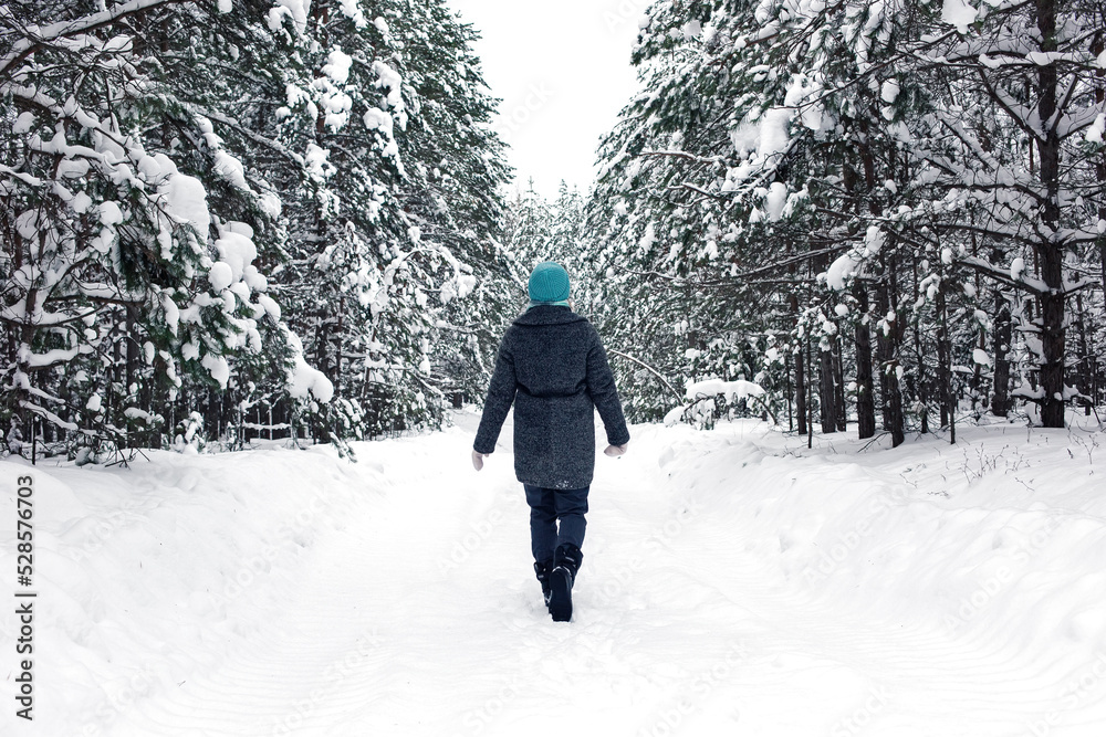 A lonely girl in a short coat and a blue hat walks through a beautiful winter snowy forest