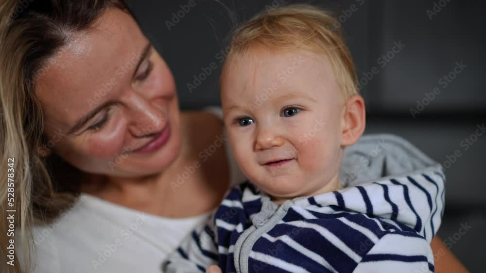 Close-up cute blond baby girl with grey eyes looking at camera smiling as woman kissing child on cheek. Portrait of curios positive happy daughter and loving mother indoors at home Close-up cute blond baby girl with grey eyes looking at camera smiling as woman kissing child on cheek. Portrait of curios positive happy daughter and loving mother indoors at home