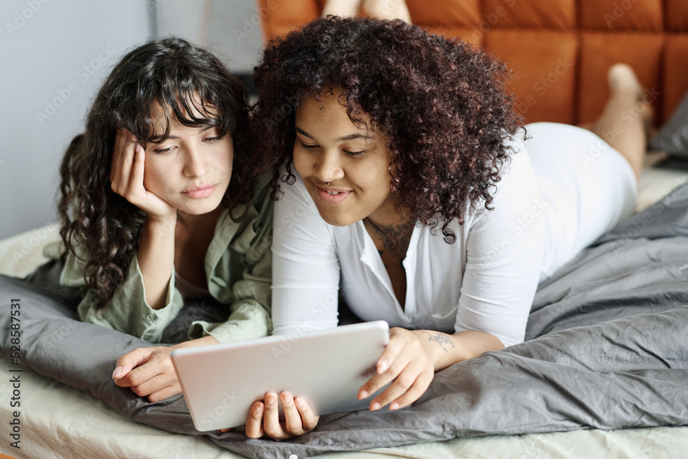 Happy young lesbian couple in home wear watching movie or choosing goods in online shop while resting on comfortable double bed Happy young lesbian couple in home wear watching movie or choosing goods in online shop while resting on comfortable double bed