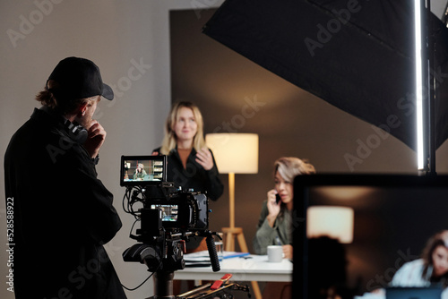 Back view of young videographer standing in front of steadicam during shooting of commercial with female model in studio
