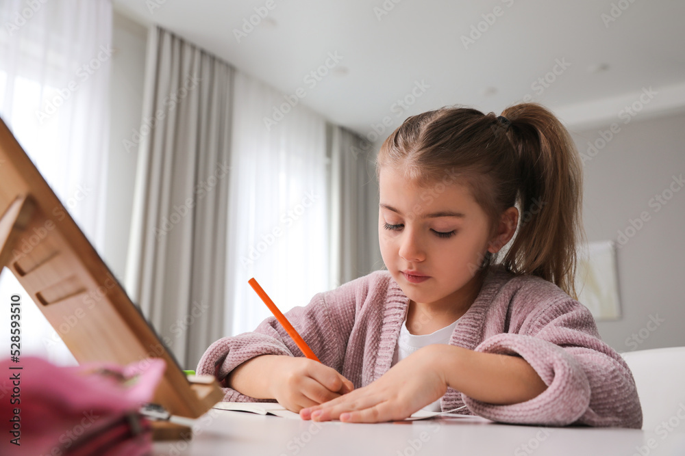 Adorable little girl doing homework with tablet at table indoors Adorable little girl doing homework with tablet at table indoors