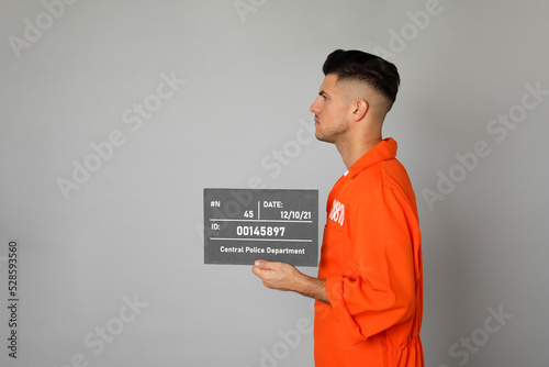 Mug shot of prisoner in orange uniform with board on grey background, side view